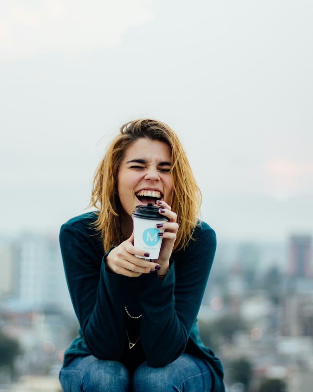A woman laughing on a rooftop, holding a coffee cup