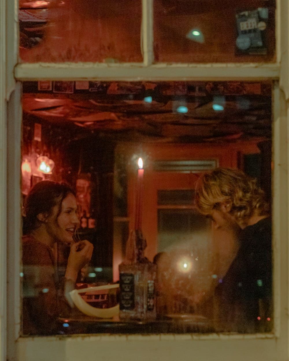 Two friends talking across a table, seen through a lit window